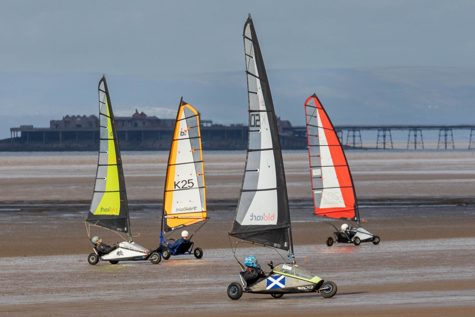 Several land yatchers racing along a sandy beach in their three-wheeled karts with colourful sails